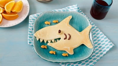 A pancake in the shape of a shark with five goldfish beside it