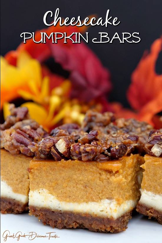Pecan-topped cheesecake pumpkin bars clustered together on a white surface with autumn leaves in the background.