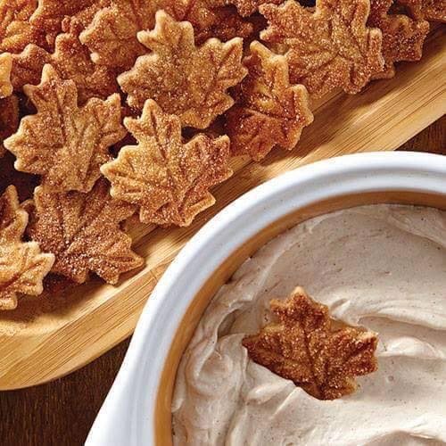 A wooden tray of Pie Crust Leaf Chips, with a bowl of creamy Cinnamon Dip right next to it. One cinnamon leaf chip is partially dunked in the creamy dip.