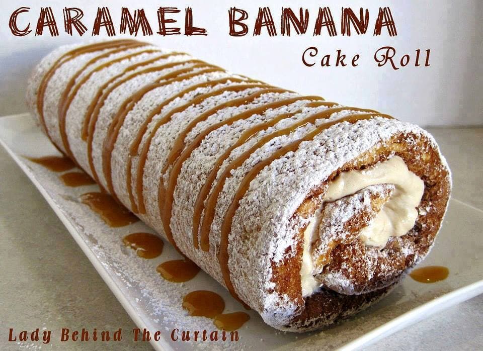 An unsliced caramel banana cake roll on a long white platter. The cake roll is dusted generously with powdered sugar, and iced horizontal lines of caramel drizzle.