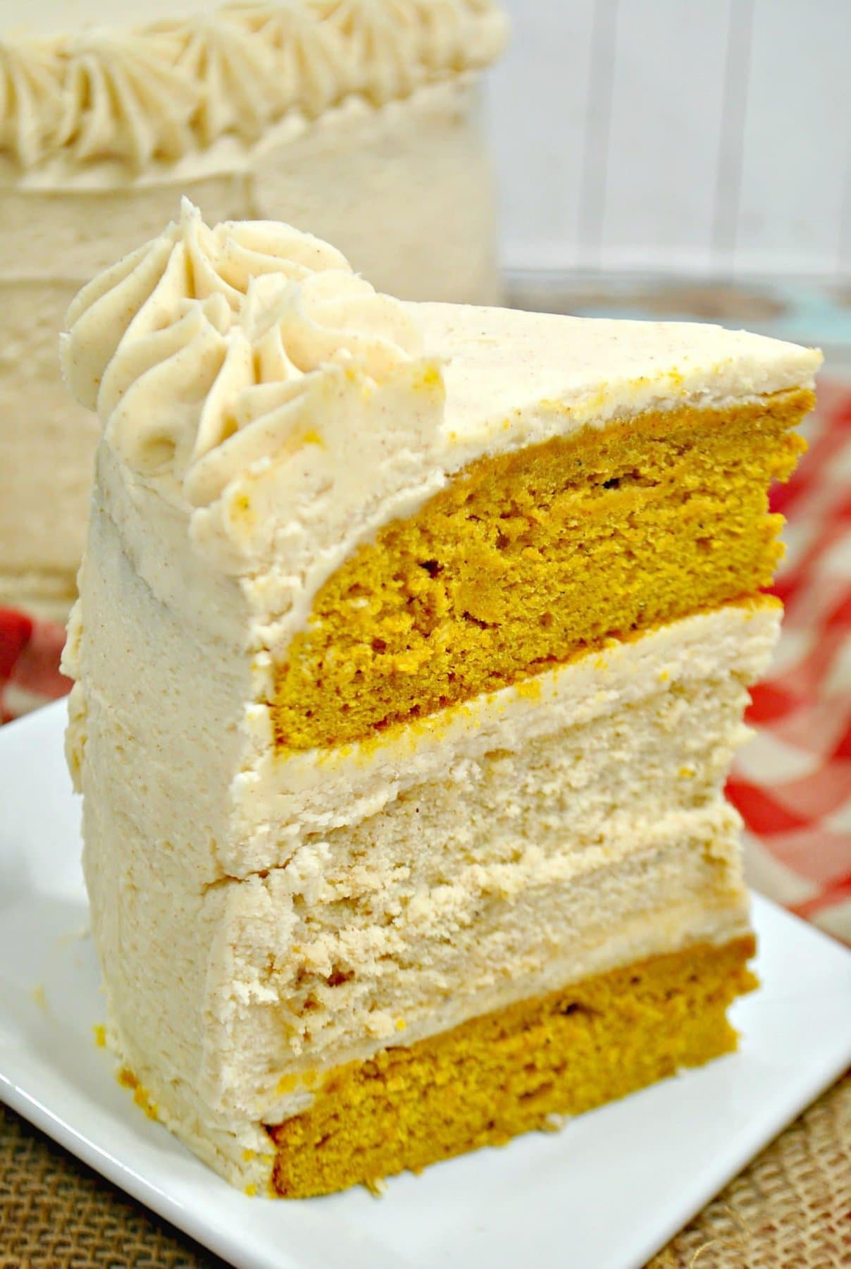 A tall, thick slice of layered pumpkin cheesecake cake, frosted with cinnamon-cream-cheese buttercream-style frosting, on a white plate. The table is covered in burlap and gingham.