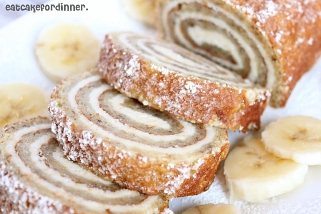 Sliced banana cake roll on a work surface dusted with powdered sugar and scattered with slices of fresh banana.
