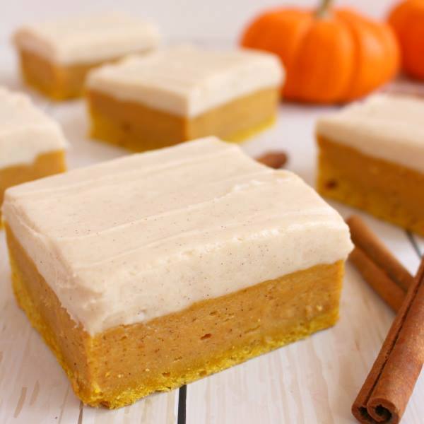 A square of pumpkin spice sugar cookie bars with cream cheese frosting on a whitewashed wooden surface with cinnamon sticks