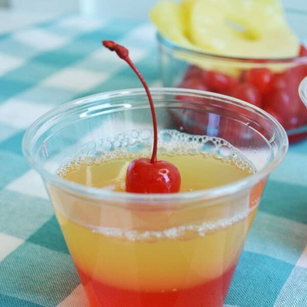 A layered jello shot with cherry jello on the bottom and pineapple jello above it with a cherry on top