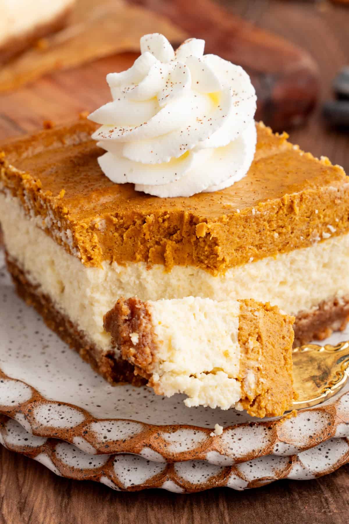These Pumpkin Cheesecake Bars is the ultimate Fall dessert that starts with a cinnamon graham cracker crust that's layered with creamy vanilla cheesecake and then topped with spiced pumpkin cheesecake. pumpkin cheesecake bar on a plate with a bite taken off with a fork