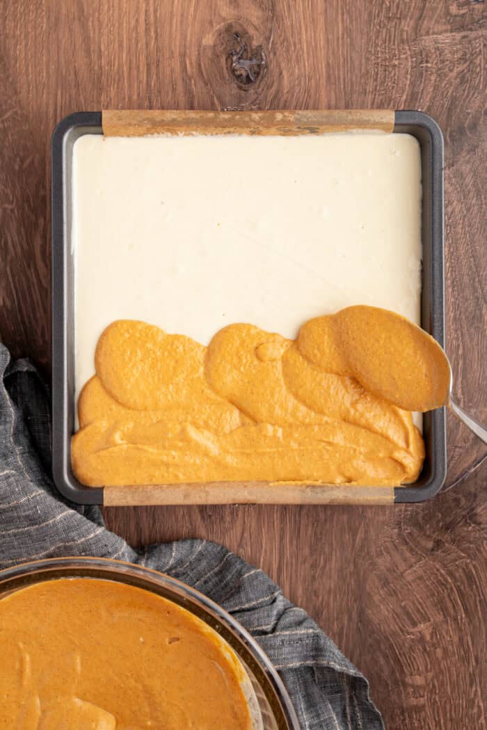 These Pumpkin Cheesecake Bars is the ultimate Fall dessert that starts with a cinnamon graham cracker crust that's layered with creamy vanilla cheesecake and then topped with spiced pumpkin cheesecake. pumpkin cheesecake mixture being spread over the vanilla cheesecake layer in a pan