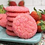 One cookie leaning against a stack of strawberry champagne cookies on a blue plate, with fresh berries in the background.