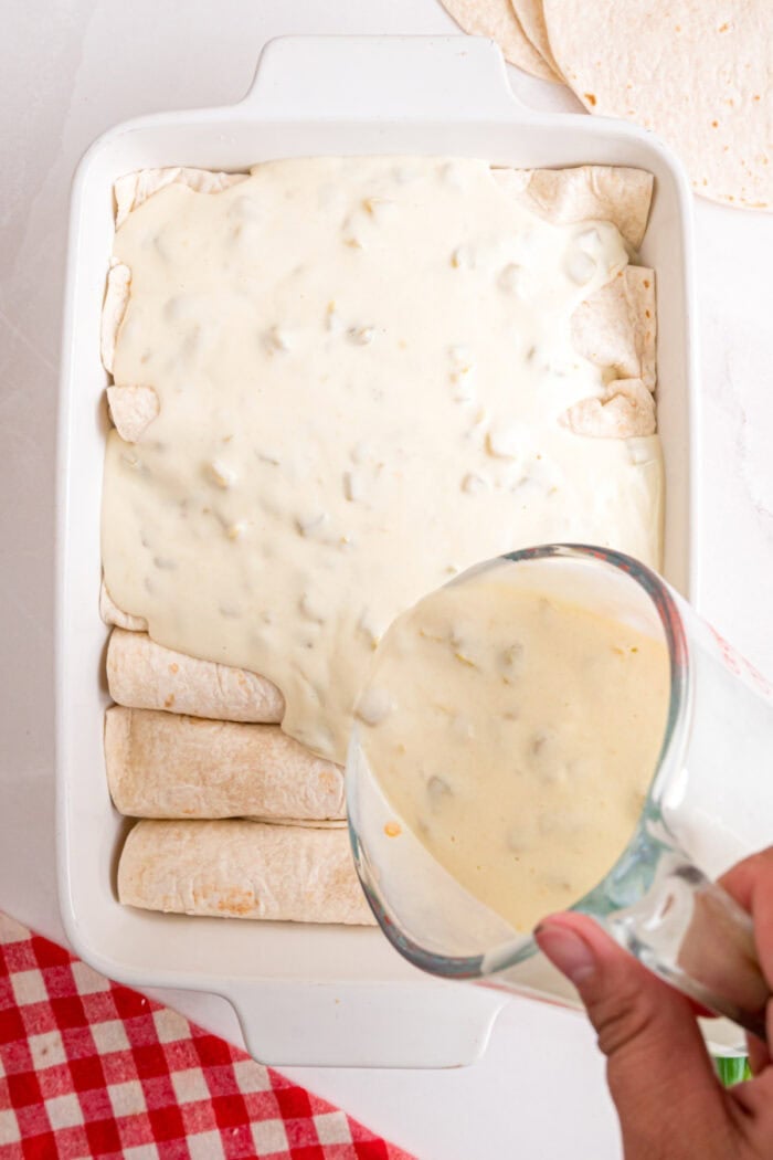 These White Chicken Enchiladas are made with rotisserie chicken, shredded cheese, and a simple sauce made with sour cream and green chiles. pouring white sauce over chicken enchiladas