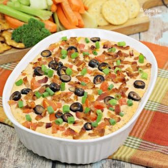 Warm bacon cheddar dip topped with olives, green onions, and bacon in an oval baking dish, with a platter of crackers and veggies in the background.