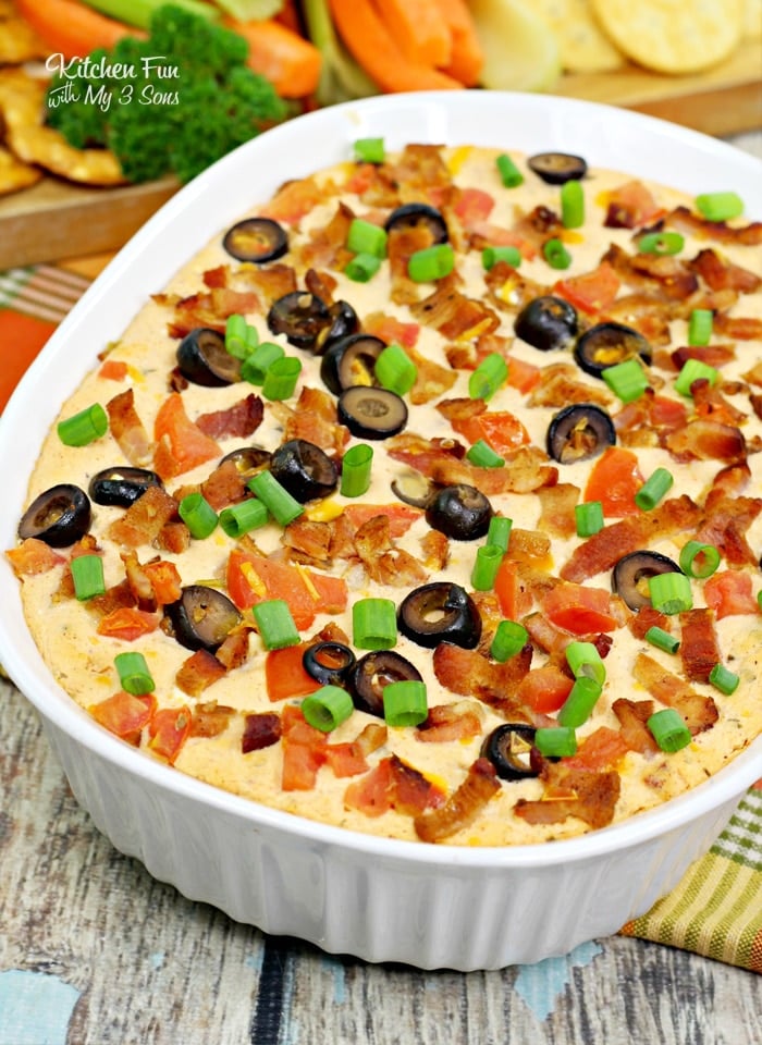 Warm bacon cheddar dip topped with olives, green onions, and bacon in an oval baking dish, with a platter of crackers and veggies in the background.