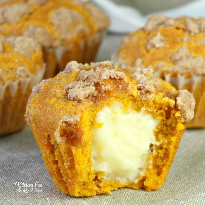 Three pumpkin cream cheese muffins on a beige tabletop. The muffin in the foreground has been cut in half to reveal the cream cheese filling. 