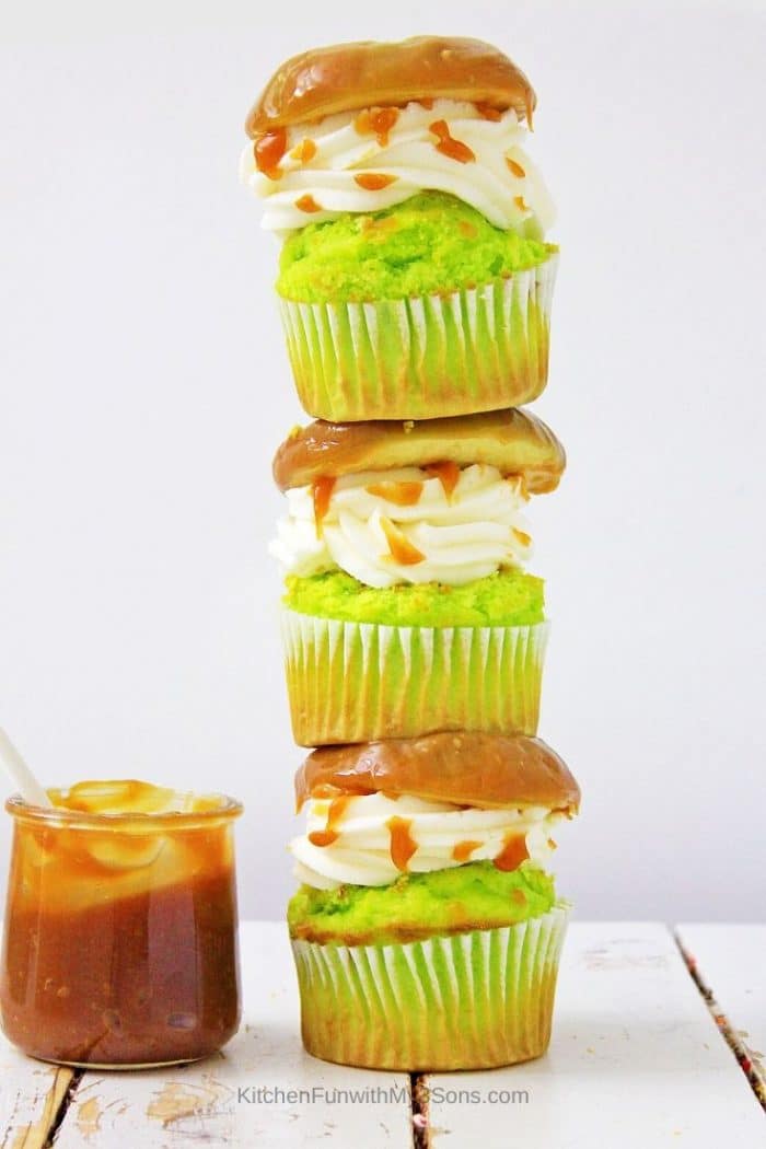 A stack of three caramel apple cupcakes on a white wooden surface with jar of caramel on side