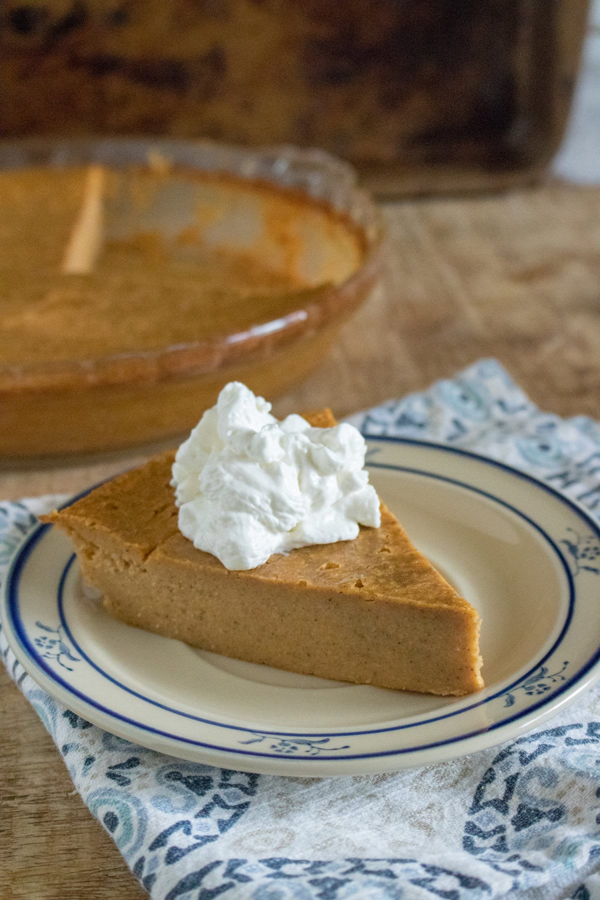 A slice of Crustless Pumpkin Pie topped with whipped cream on a plate, next to the rest of the pie on a wooden table.