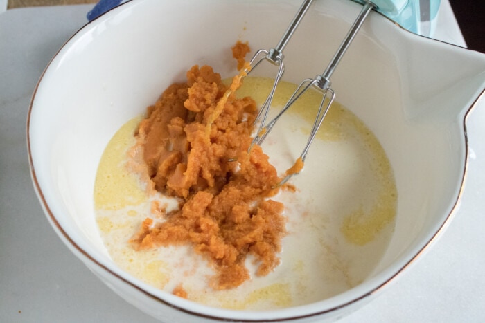 Pumpkin puree added to the bowl of a stand mixer with pumpkin pie batter ingredients.