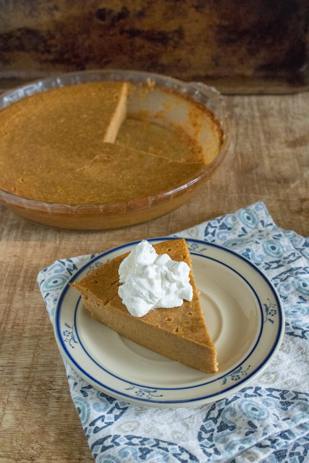 A slice of Crustless Pumpkin Pie topped with whipped cream on a plate, next to the rest of the pie on a wooden table.