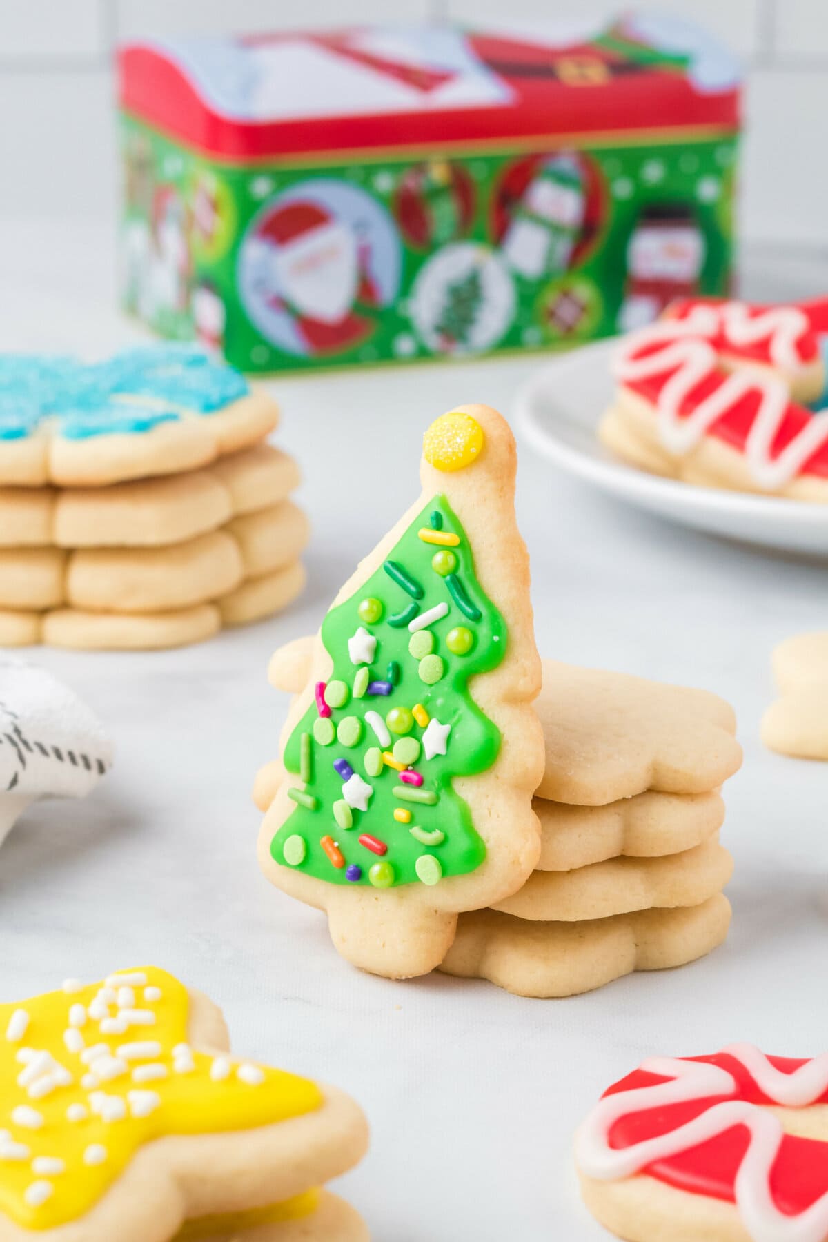 A cut out sugar cookie Christmas tree leaning against another stack of cookies