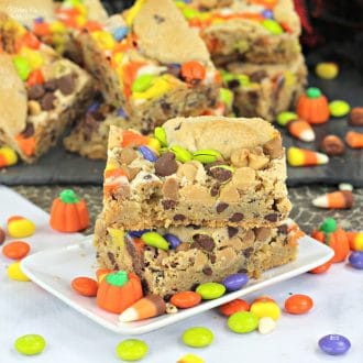 Two Halloween candy cookie bars stacked on a plate surrounded by scattered candy, with more cookie bars in the background.