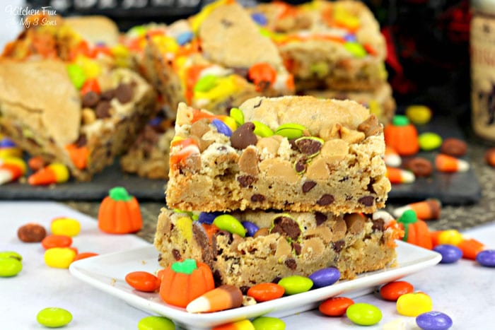 Two Halloween candy cookie bars stacked on a plate surrounded by scattered candy, with more cookie bars in the background.