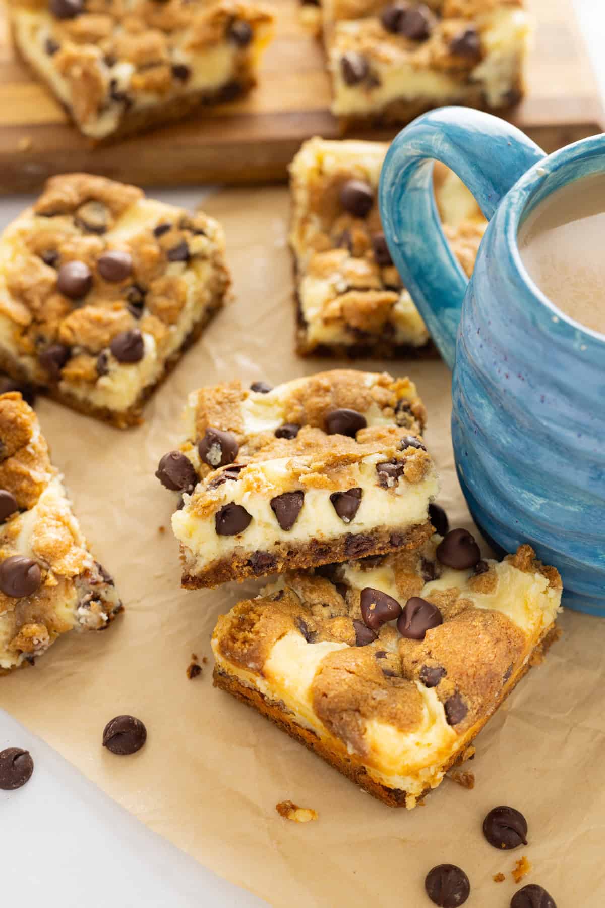 Creamy, tangy cheesecake filling is stuffed between layers of cookie dough in these Chocolate Chip Cheesecake Bars. Chocolate chip cheesecake bars on parchment paper next to a mug