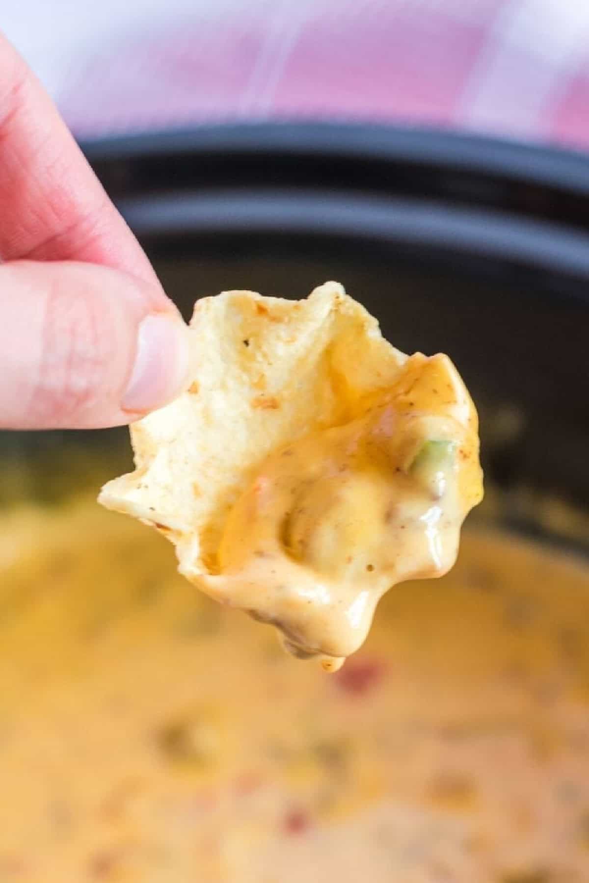 Velveeta cheese, Rotel, and ground beef come together in this cheesy and flavorful Hamburger Dip. Made in the slow cooker! A hand holding a tortilla chip over a crockpot of cheesy hamburger dip