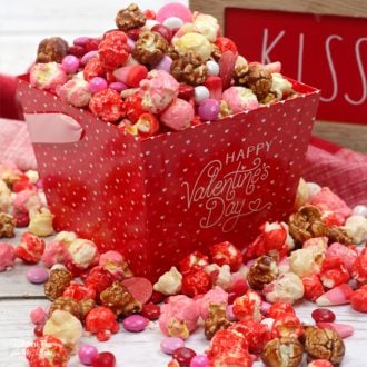 A red container of Valentine popcorn party mix with popcorn scattered over a table.