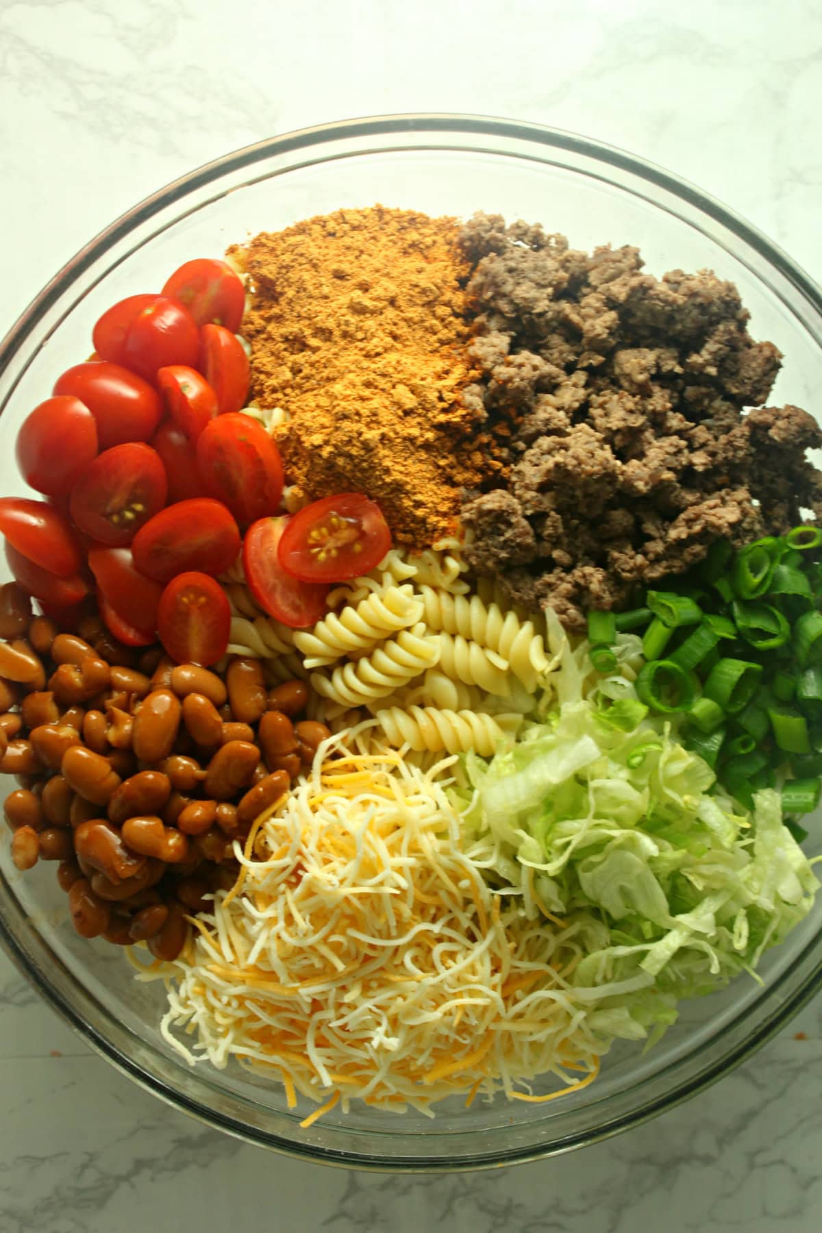 A fun twist on a classic, Taco Pasta Salad is made with rotini, ground beef, and your favorite taco toppings tossed with French dressing. taco pasta salad ingredients in a bowl