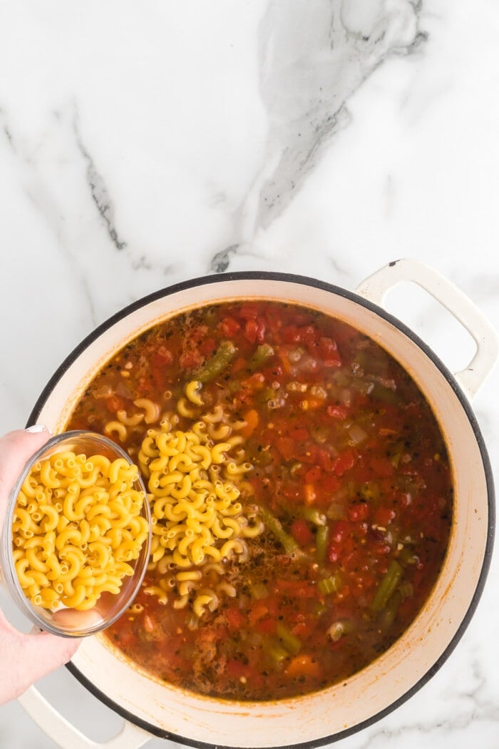 Elbow macaroni being added to a pot of minestrone soup