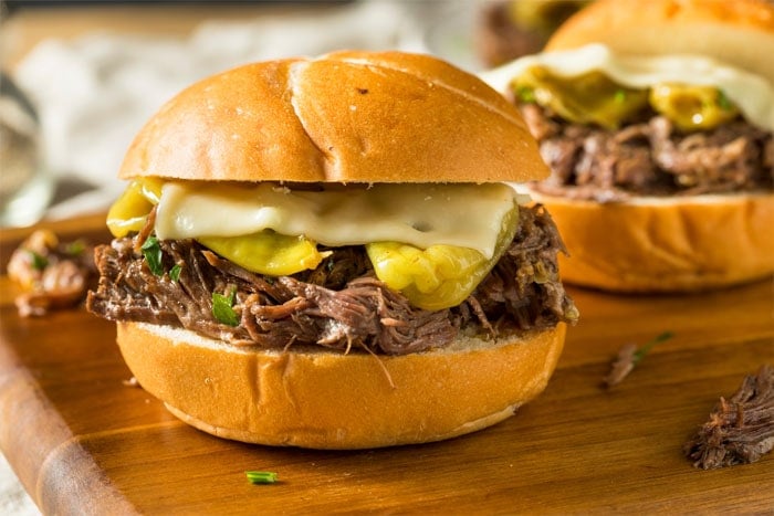 A shredded beef sandwich on a large sweet roll, topped with pepperoncini and melted Swiss cheese.