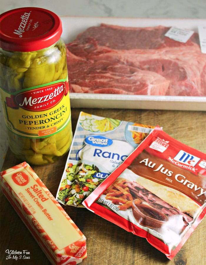 From top: A large chuck roast in its packaging, a packet of au jus gravy mix, a packet of ranch dressing seasoning, a stick of butter, and a jar of pepperoncini.