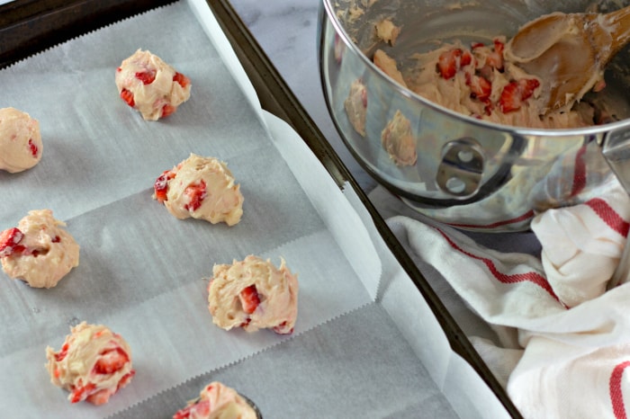 White chocolate strawberry cookie dough balls on a lined baking sheet, next to cookie dough in a metal mixing bowl.