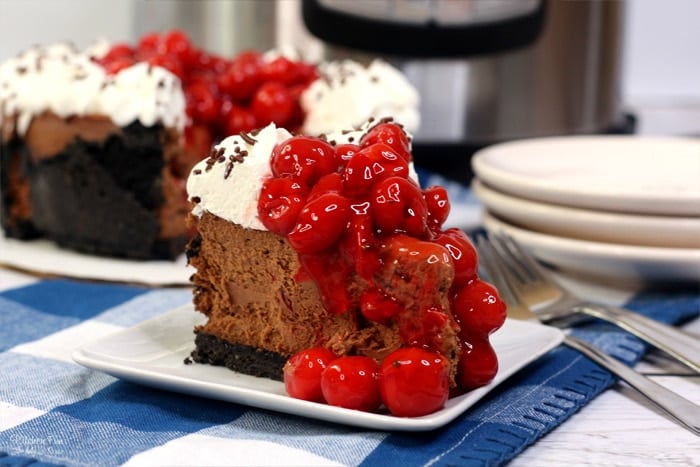 A slice of black forest cheesecake on a plate topped with swirls of whipped cream and cherries.