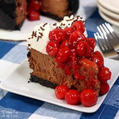 A slice of black forest cheesecake on a plate topped with swirls of whipped cream and cherries.