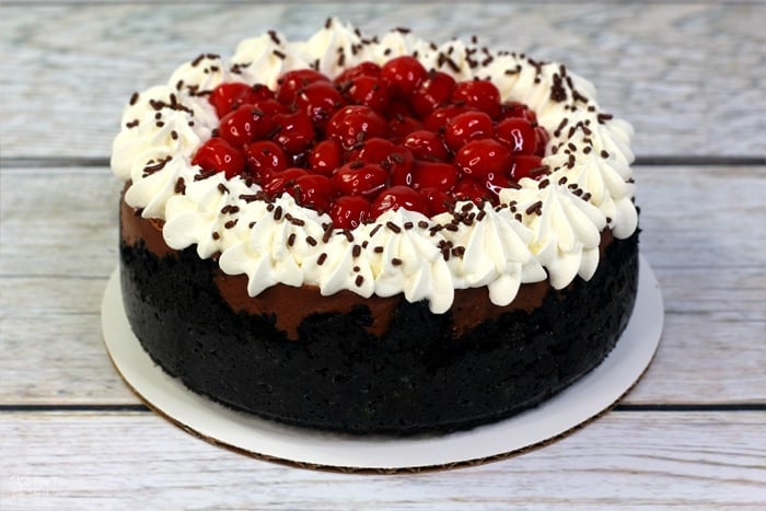 A whole black forest cheesecake topped with whipped cream swirls with cherries in the middle.