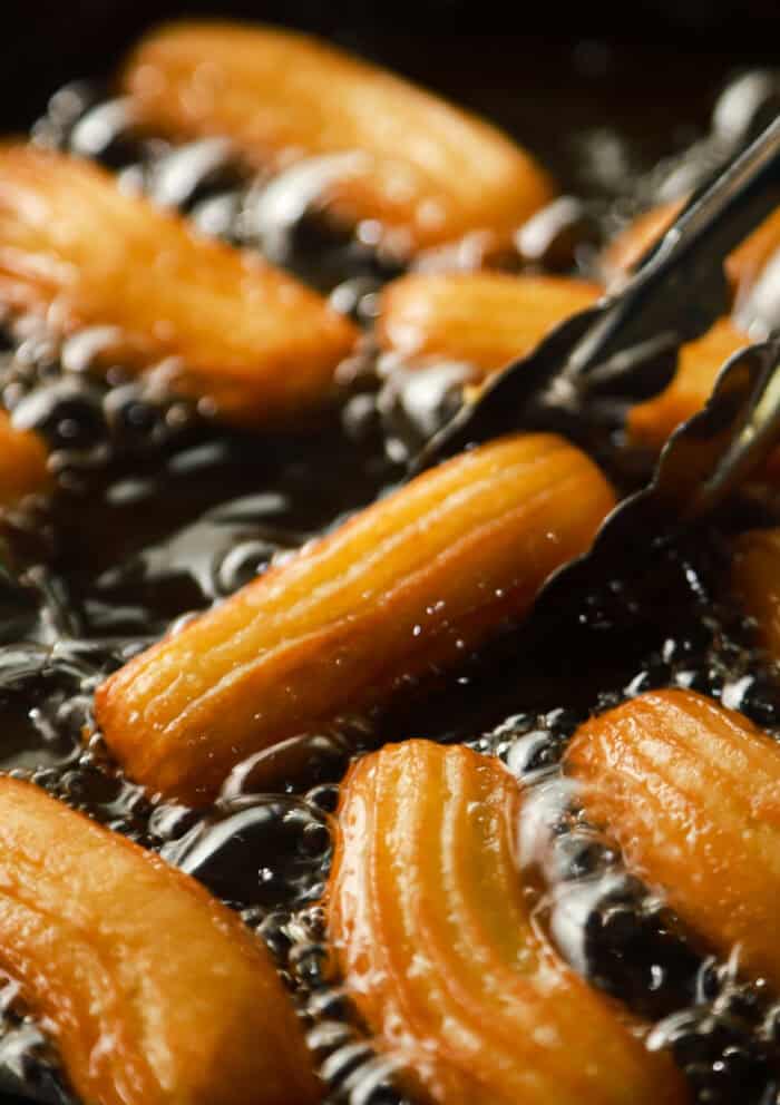 These Churros are soft on the inside, crisp and golden brown on the outside, and tossed with cinnamon sugar. Tongs removing a churro from oil