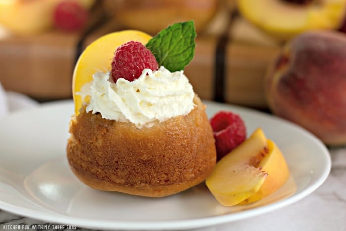 A mini peach upside down cake topped with whipped cream, a raspberry, a peach slice and a mint leaf