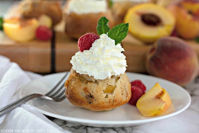 A mini peach cake on a plate with a fork, a few fresh peach slices and a couple of raspberries