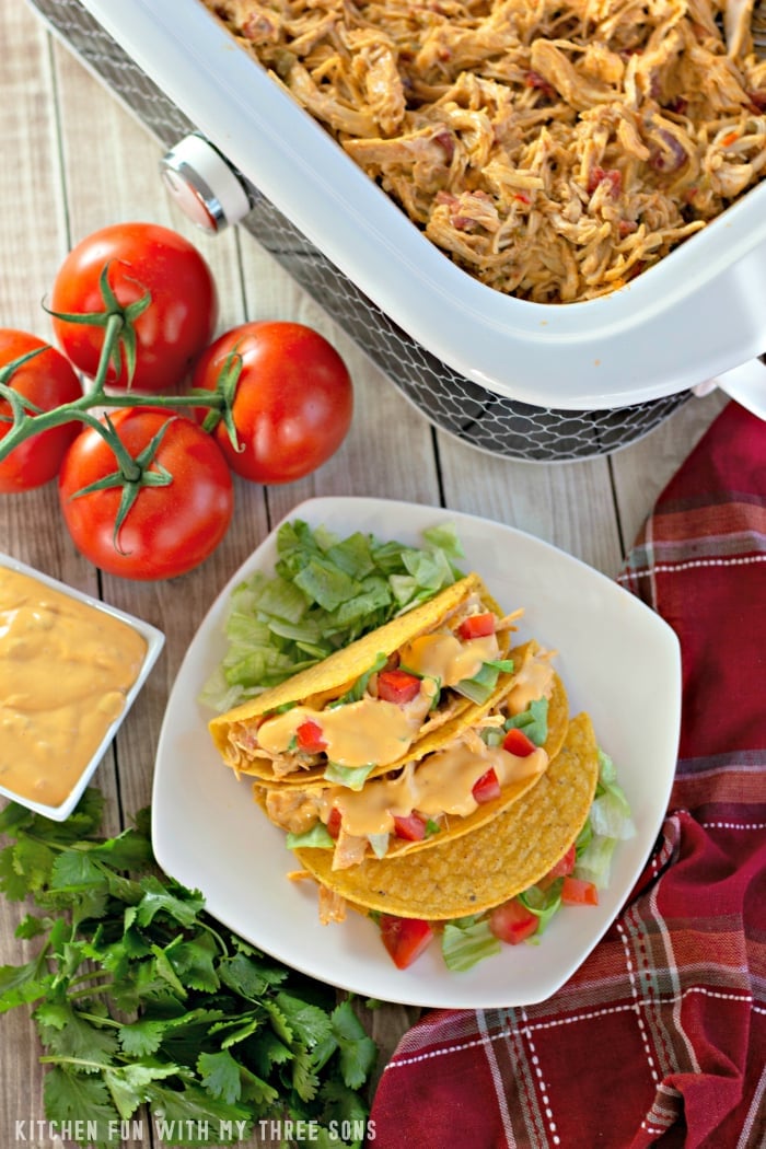 Three slow cooker chicken queso tacos on a plate beside four ripe tomatoes