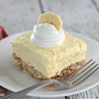 A single square of banana cream cheesecake topped with whipped cream and a banana slice.