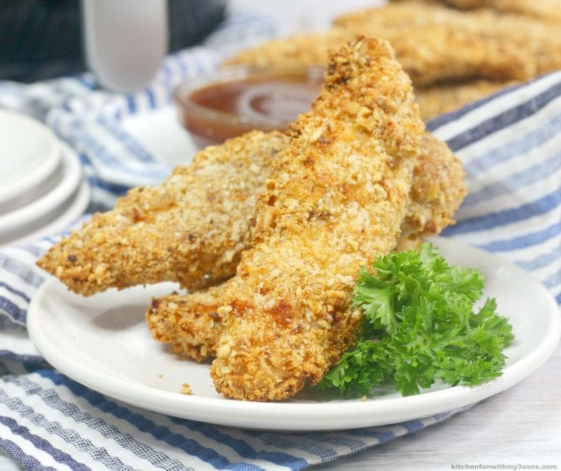 Air fryer pretzel crusted chicken tenders on a white plate next to a garnish of greens, with dipping sauce in the background.