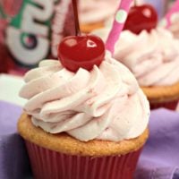 Close up of Strawberry Crush soda cupcakes topped with swirls of frosting and maraschino cherries, with cans of strawberry soda in the background.