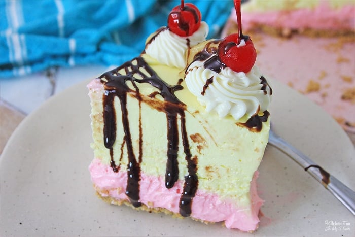 A slice of banana split cheesecake on a plate, topped with whipped cream, chocolate sauce, and maraschino cherries.