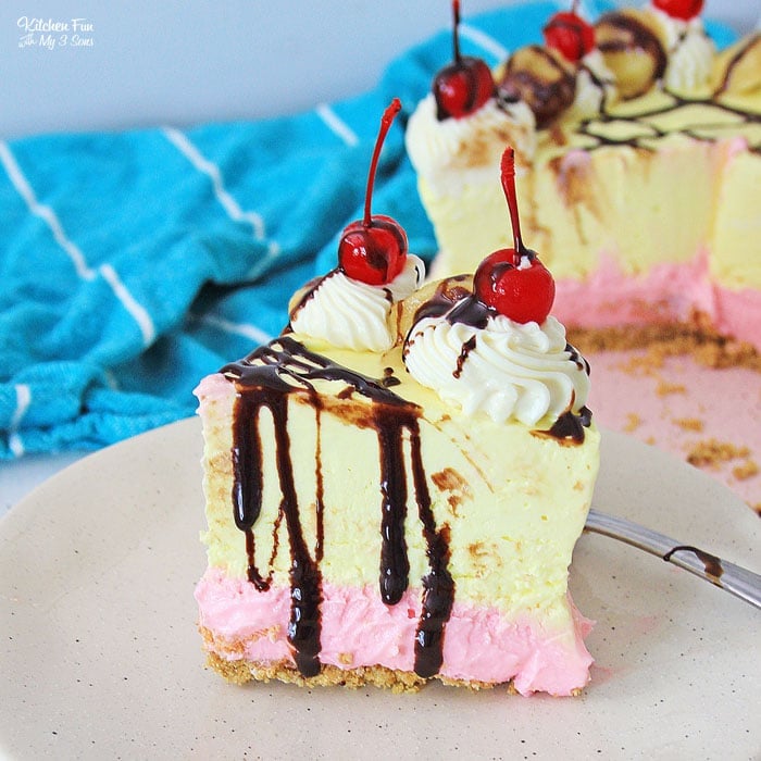 A slice of banana split cheesecake on a plate, topped with whipped cream, chocolate sauce, and maraschino cherries.