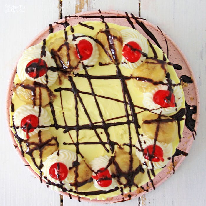 Overhead view of a banana split cheesecake decorated with a drizzle of chocolate, whipped cream, and maraschino cherries.