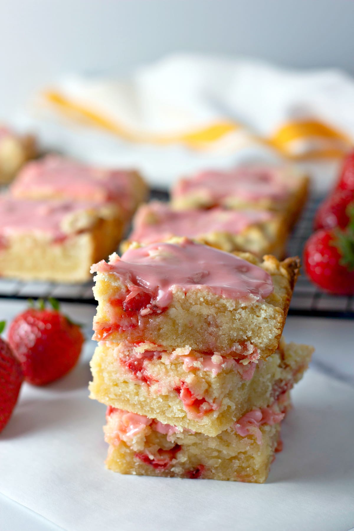 Three strawberry lemon blondies stacked on a countertop next to fresh strawberries, with more blondies in the background.