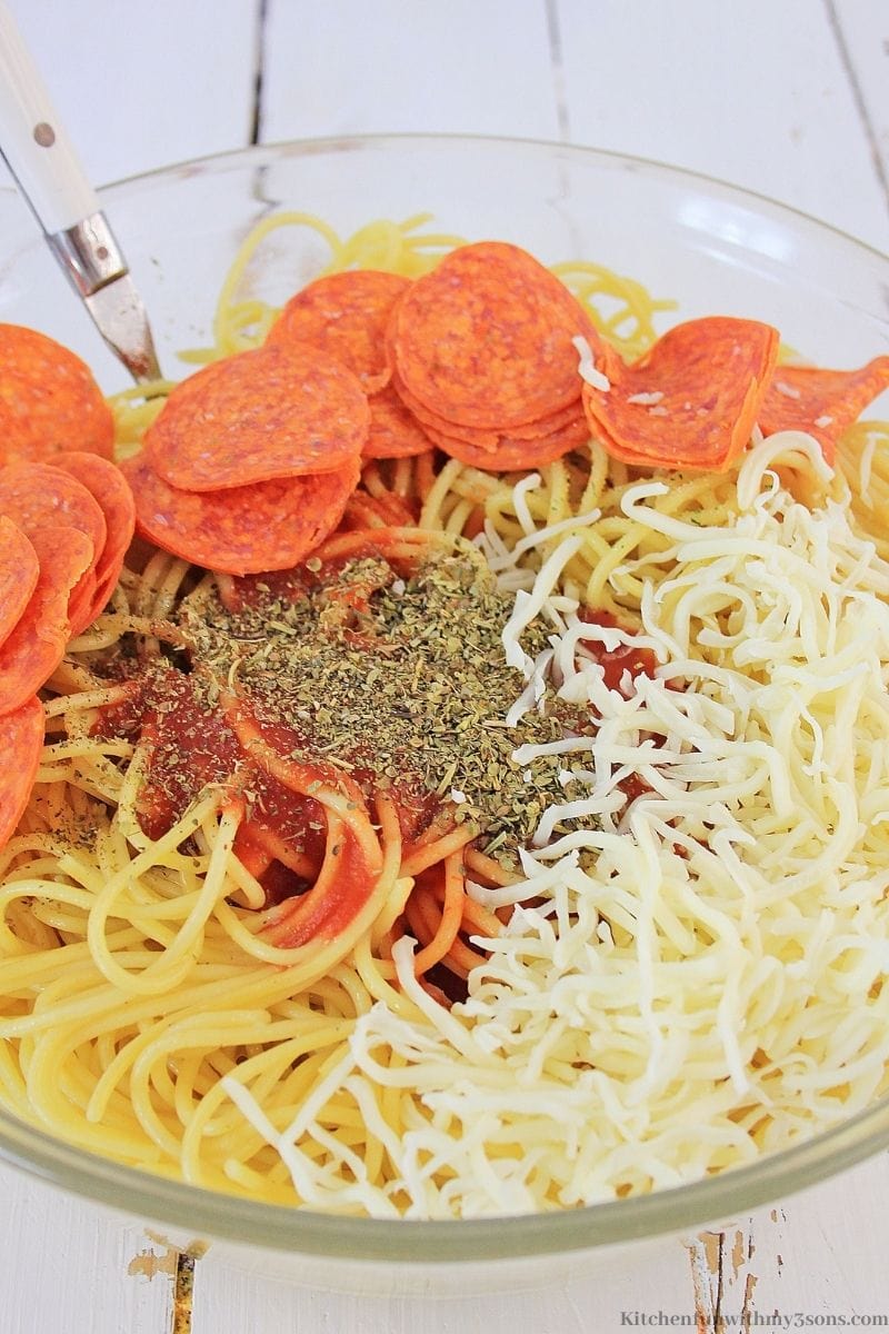 Adding pepperoni, seasoning, sauce and cheese to the pasta in a large bowl.