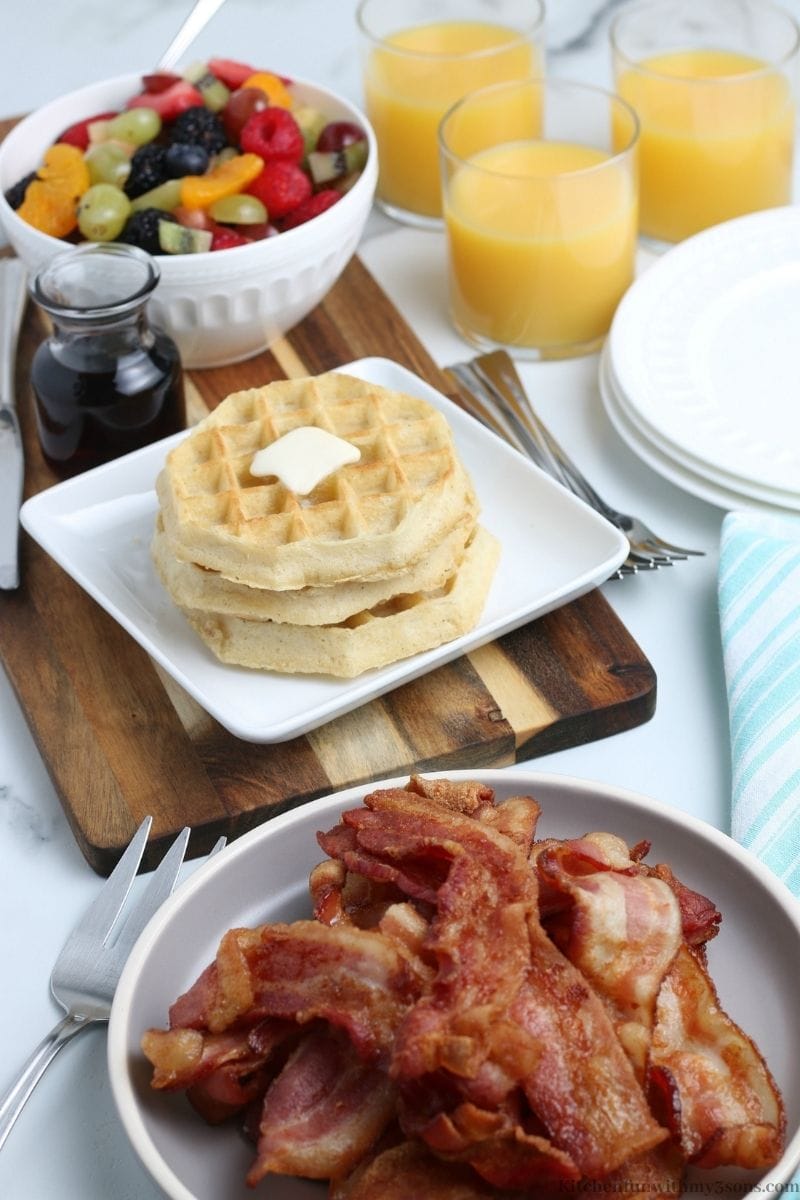 A plate of air fryer bacon next to a stack of waffles and glasses of orange juice.