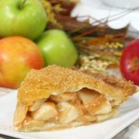 A slice of Bourbon Apple Pie laid out on a plate with a pile of whole apples in the background.