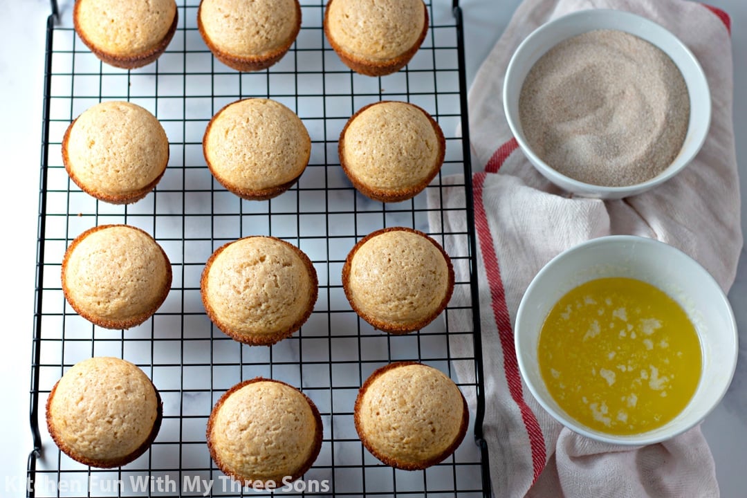 Twelve freshly-baked muffins on a cooling rack beside a bowl of melted butter and a bowl of cinnamon sugar