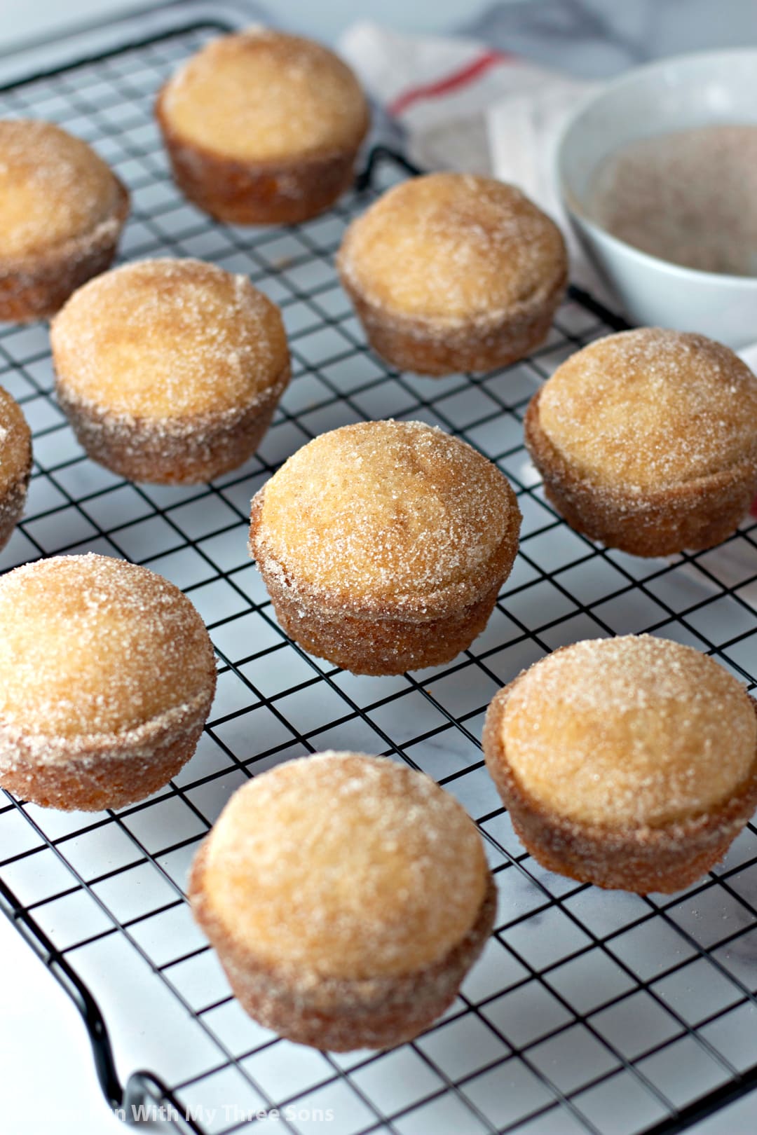 Cinnamon sugar donut muffins on a cooling rack with a bowl of cinnamon sugar behind them