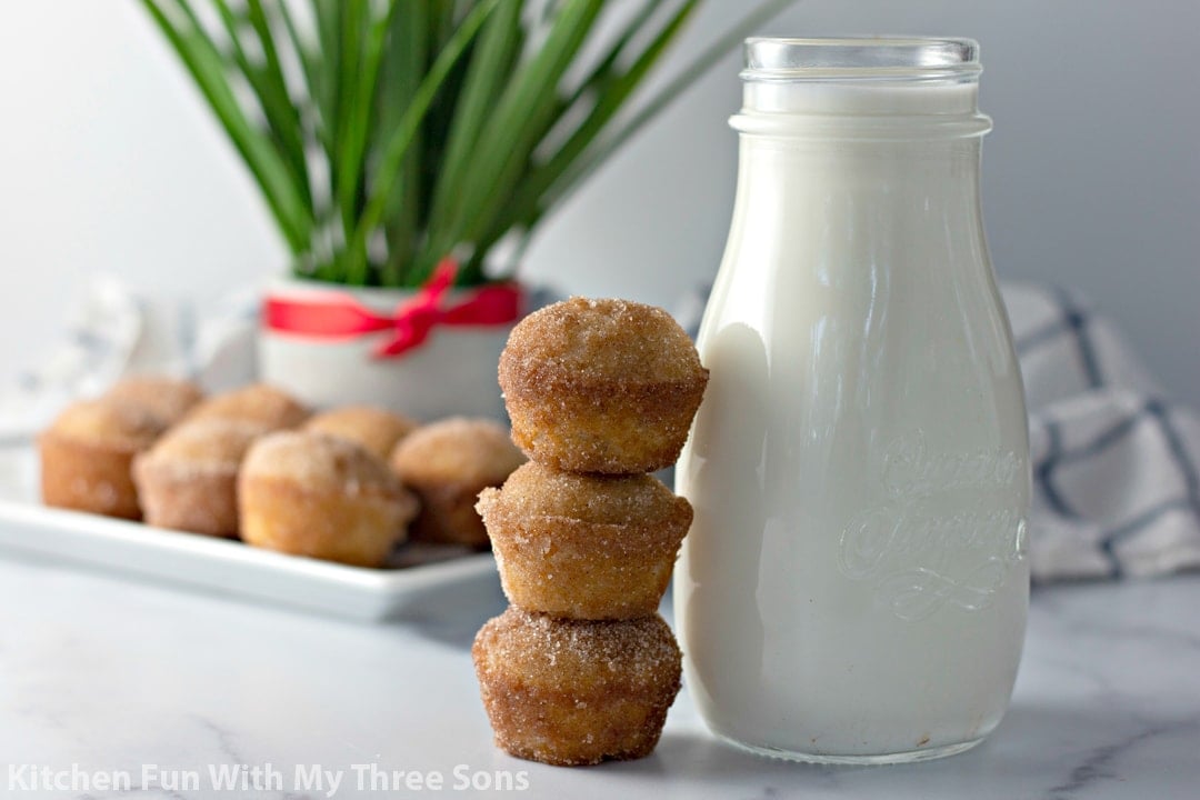 Three donut muffins stacked against a glass of milk with more muffins in the background